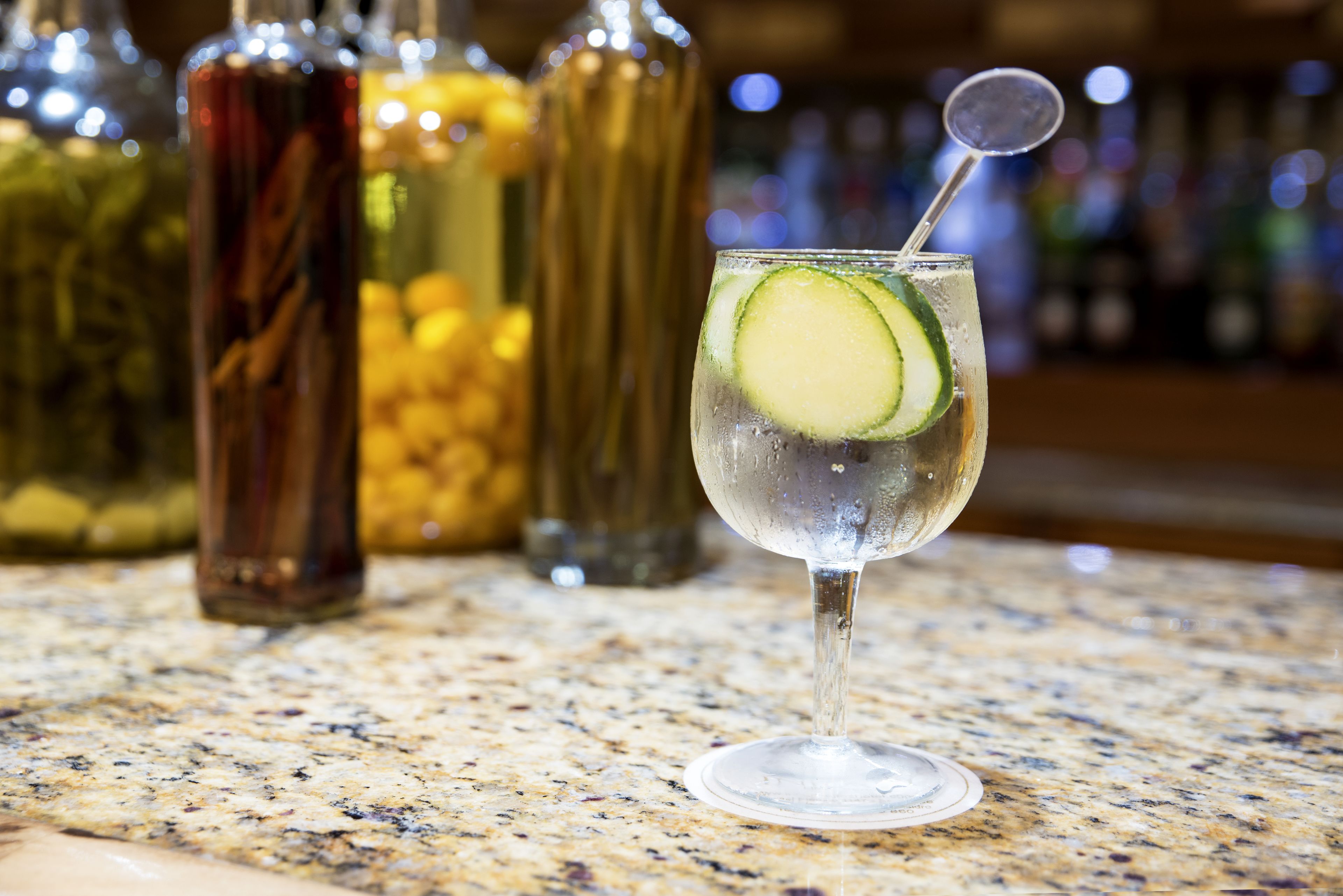 A bar setting with a glass of a clear cocktail garnished with a slice of cucumber and a round garnish on a granite countertop.