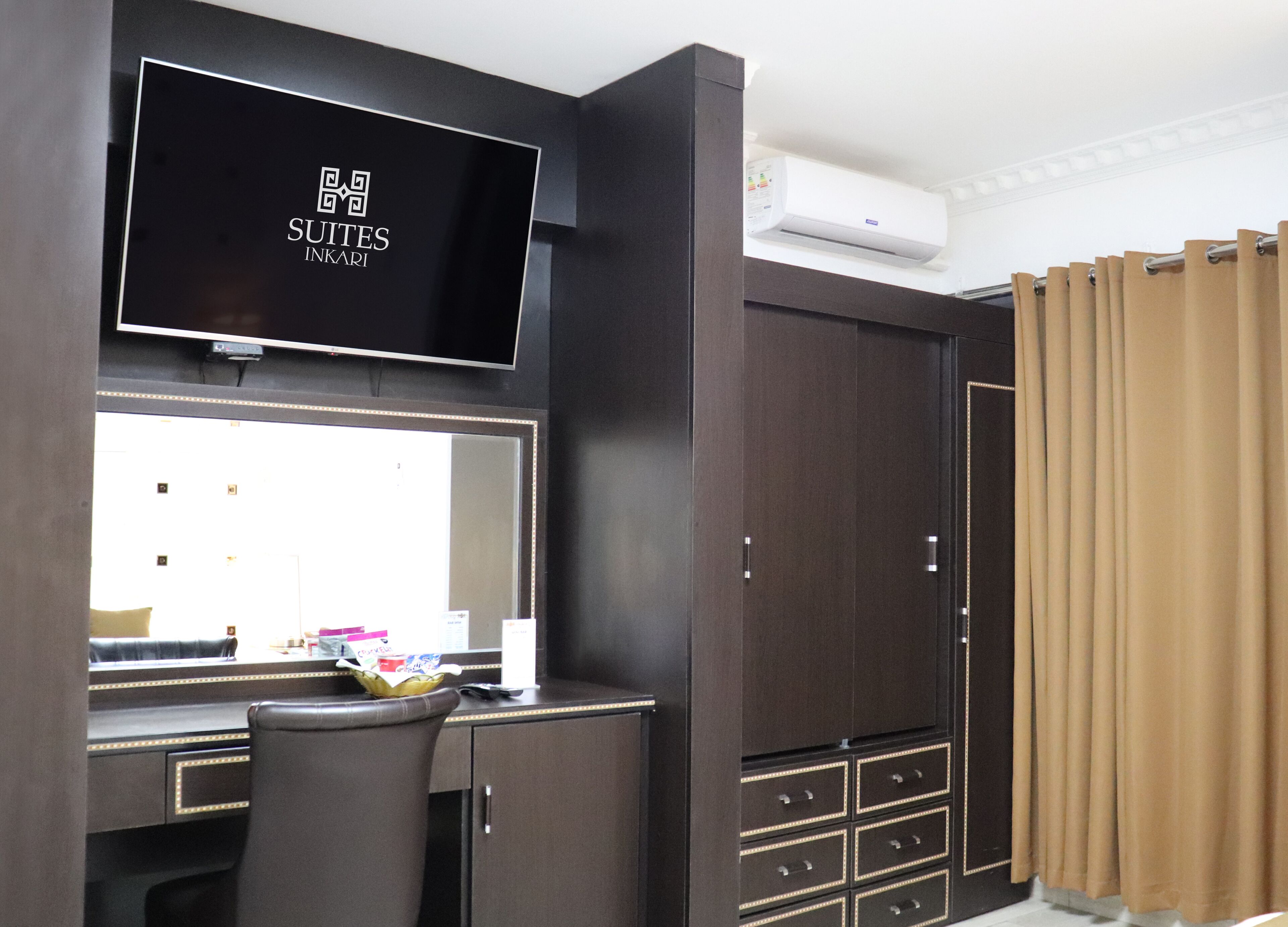A hotel room with a large dark wood wardrobe, a flat-screen TV mounted above a desk, and a large mirror.