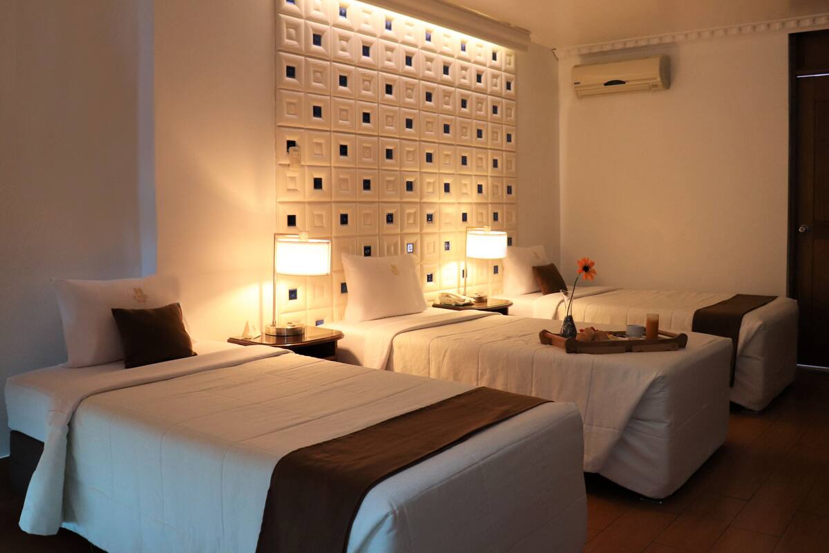 Habitación de hotel con dos camas, una pared con diseño geométrico, una mesita de noche con lámpara y un jarrón con una flor.