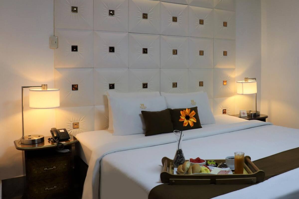 Habitación de hotel con una cama grande, dos lámparas de noche, una bandeja con alimentos para el desayuno y un cojín decorativo.