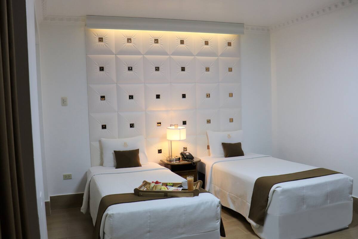 Habitación de hotel con dos camas, una mesita de noche con lámpara y una bandeja con alimentos para el desayuno.