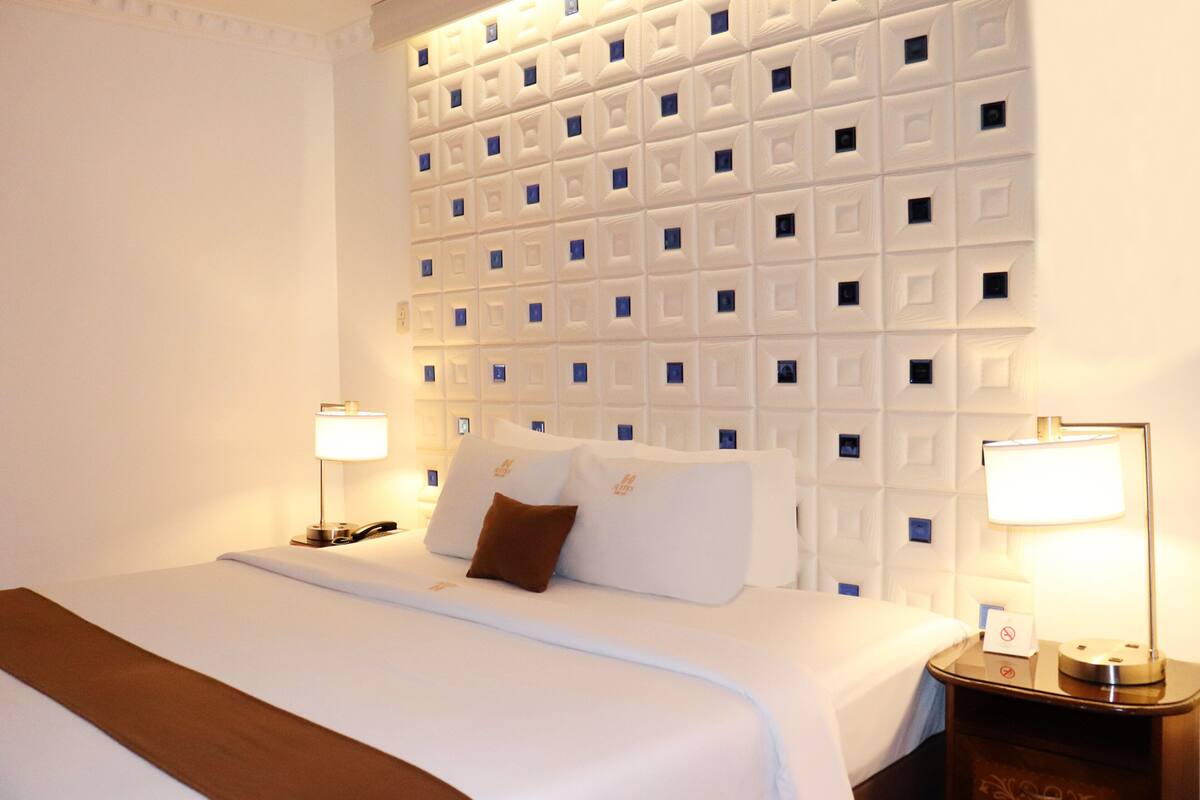Habitación de hotel con una cama grande, dos mesitas de noche con lámparas, una pared con diseño geométrico y un plaid marrón.