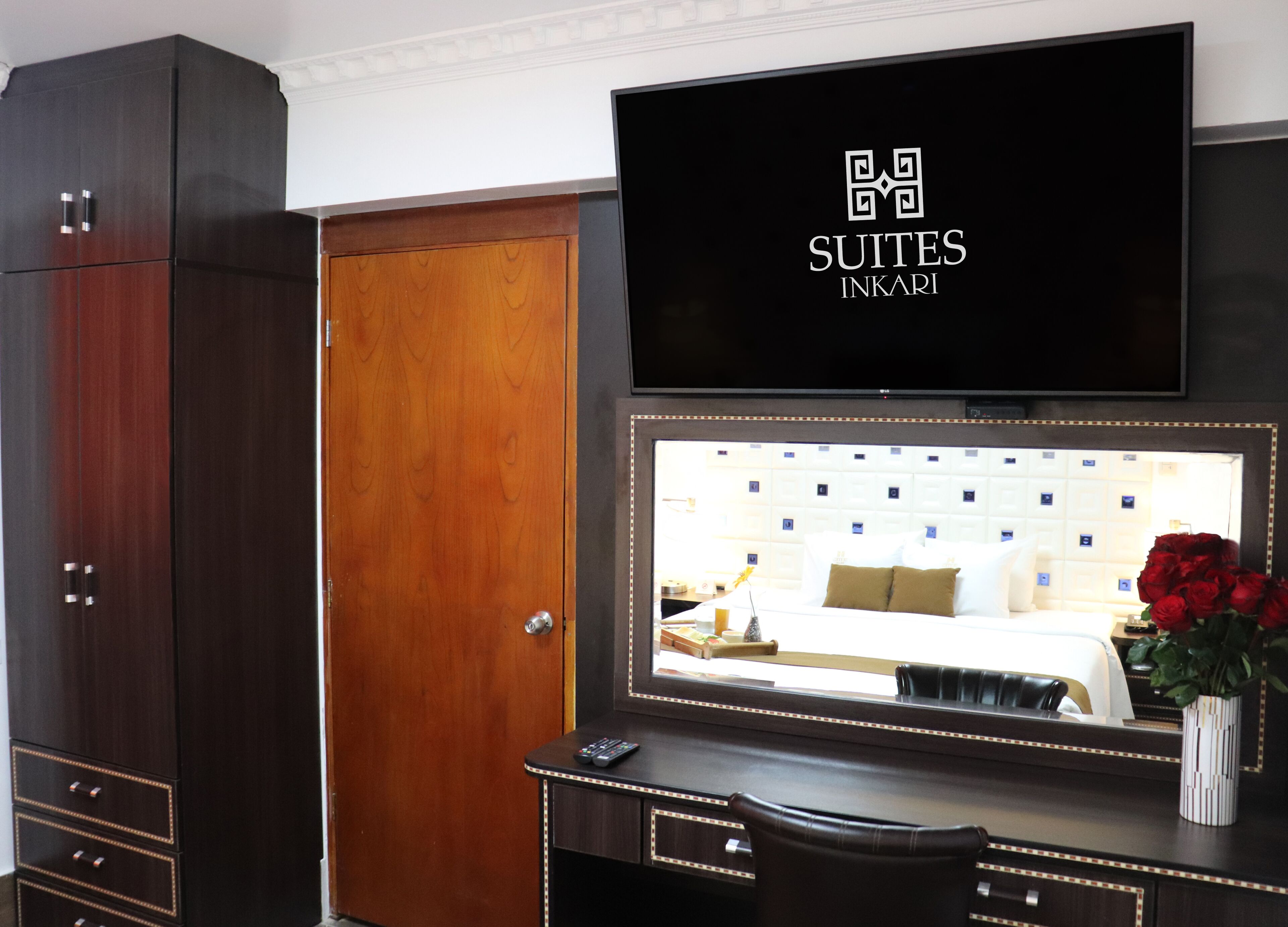 A hotel room with a large mirror reflecting a neatly made bed, a desk with a remote control, and a dark wooden wardrobe.