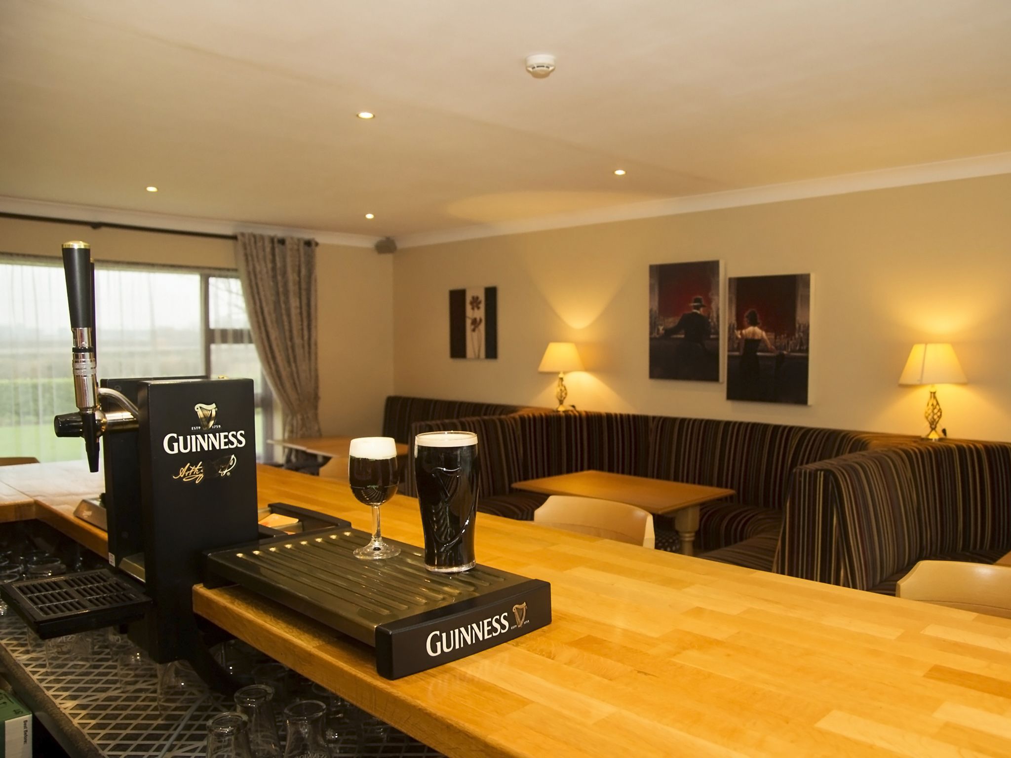 Un coin bar avec une tireuse à bière Guinness, des verres et un comptoir en bois. On aperçoit un canapé et une table au fond.