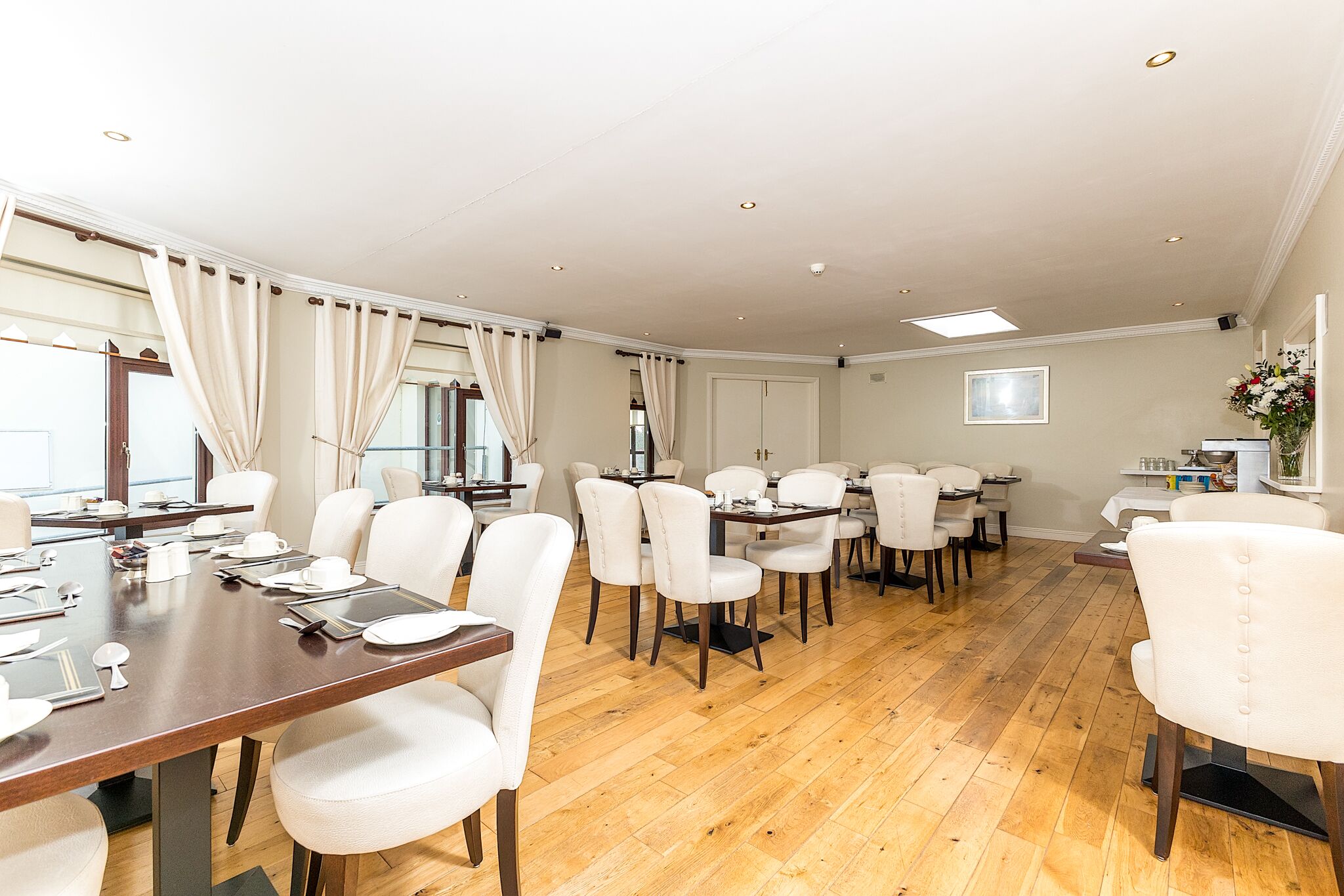 A spacious dining area with multiple tables and chairs, wooden flooring, and a large window with curtains.