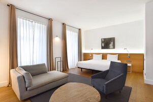 A hotel room with a bed, a sofa, a chair, a coffee table, and a painting on the wall.