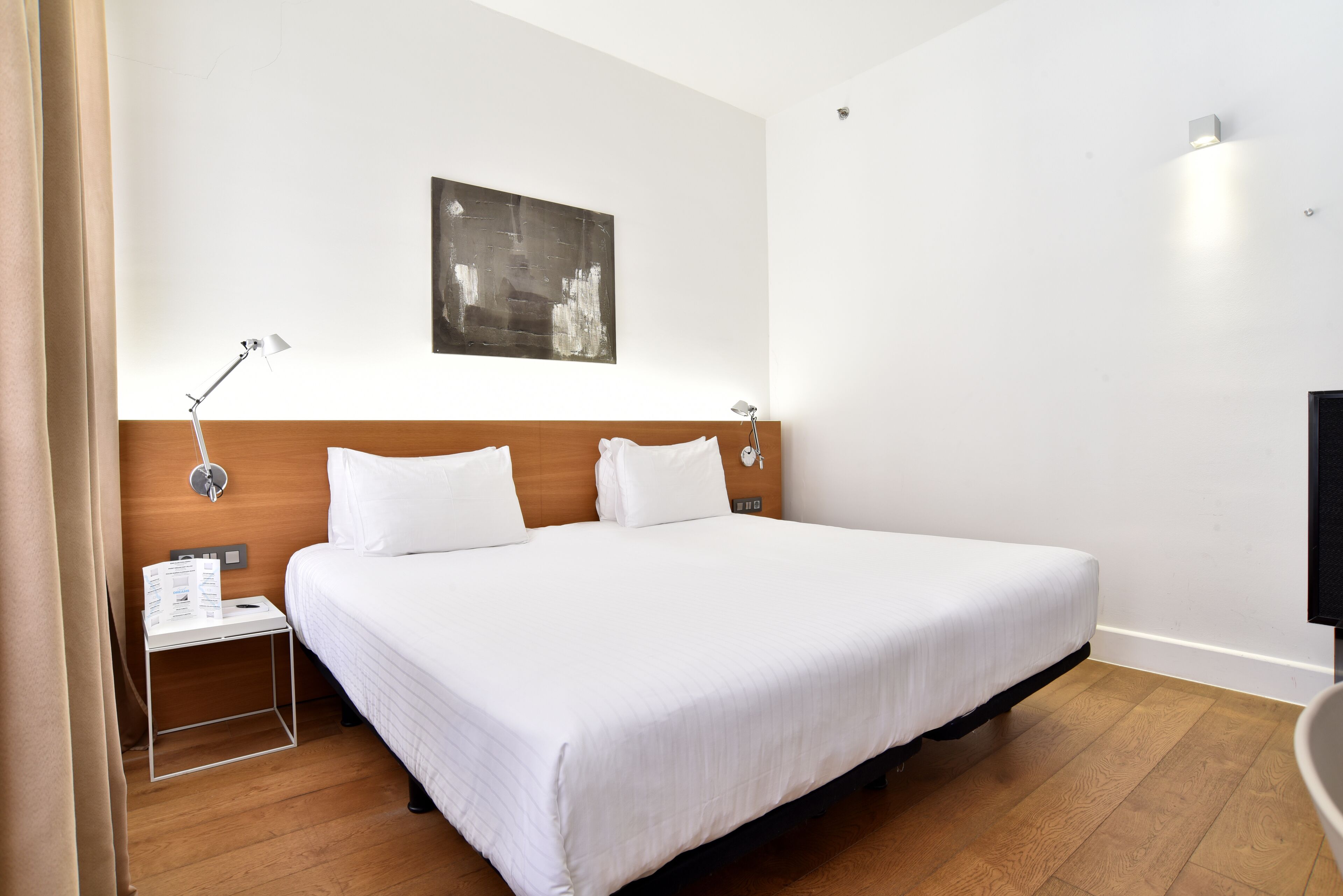 Quarto de hotel com uma cama grande, cabeceira de madeira, mesa de cabeceira com abajur e uma obra de arte na parede.