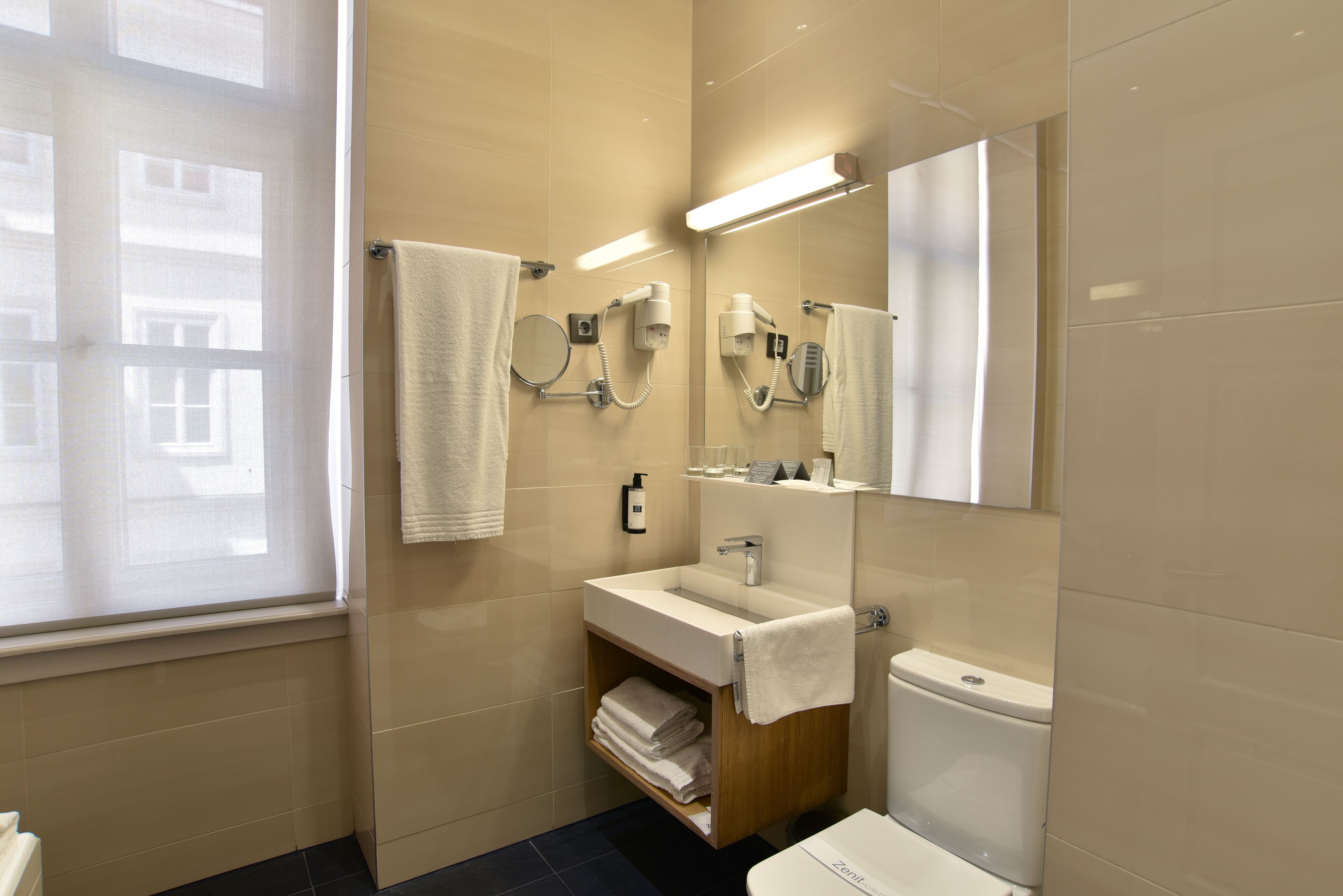 Un baño moderno con lavamanos, espejo e inodoro. Se ve una ventana con vistas a edificios.