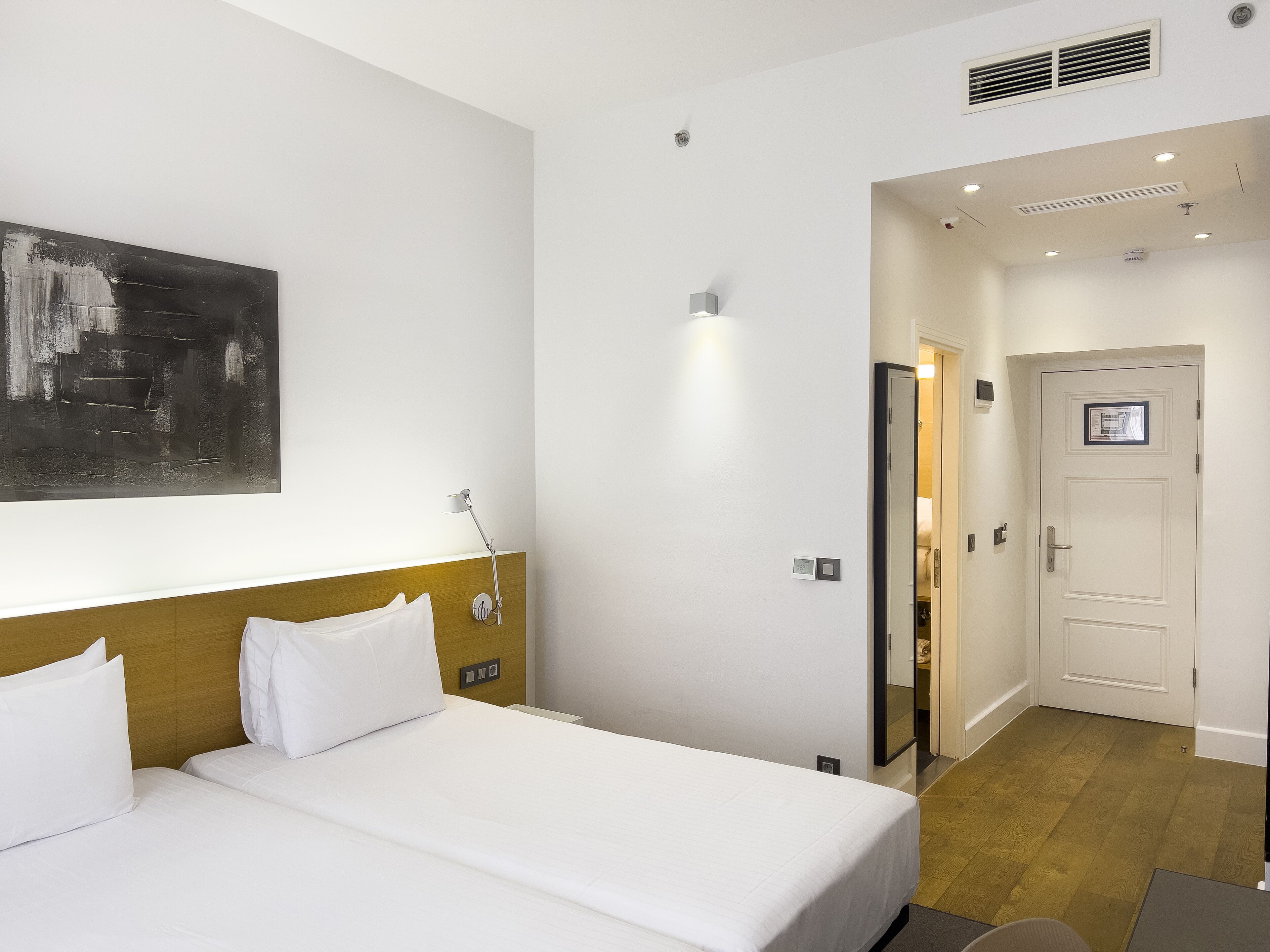 Quarto de hotel com cama, luminária de cabeceira, obra de arte na parede, porta e interruptor de luz.