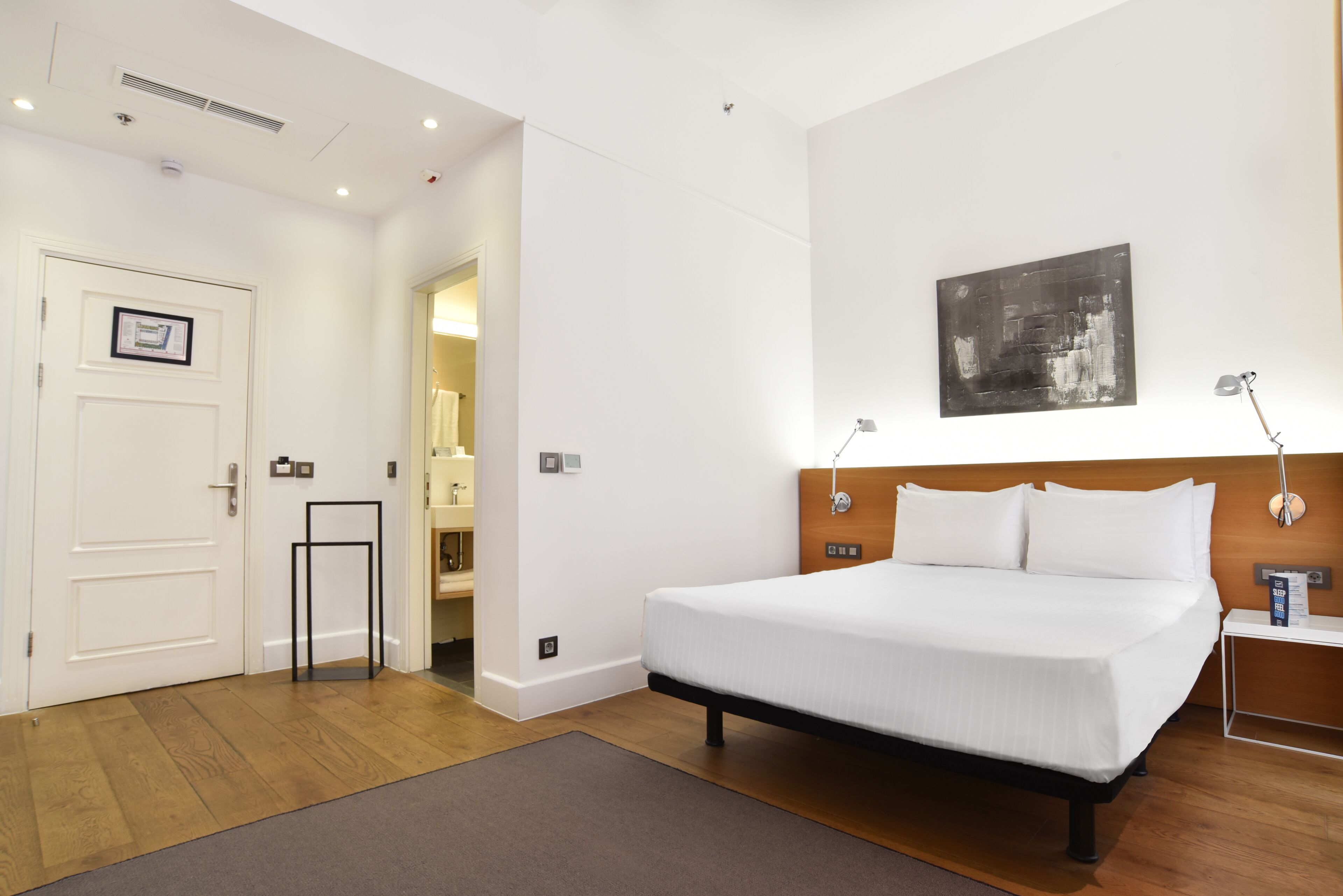 A hotel room with a large bed, a bedside table with a lamp, a framed picture on the wall, and a door leading to a bathroom.