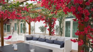 Un área de estar moderna al aire libre con un sofá blanco y cojines oscuros, rodeada de abundante buganvilla roja.