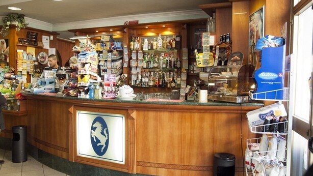 A bar area with a wooden counter, various bottles, and a sign with a horse logo. - Hotel Cavallo Bianco (Novara)