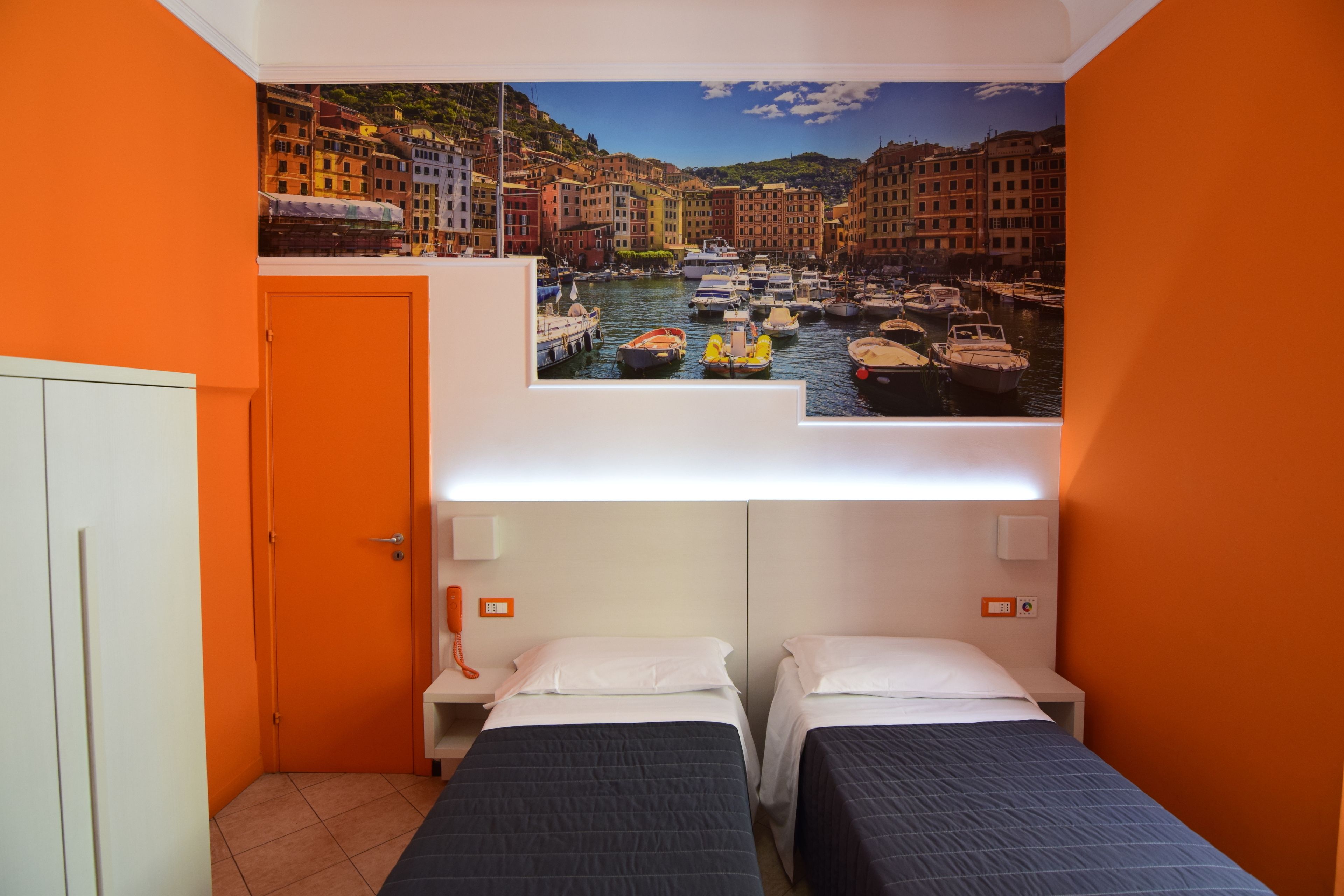 Ein Hotelzimmer mit zwei Betten, einem Hafenbild an der Wand und orangefarbenen Wänden.