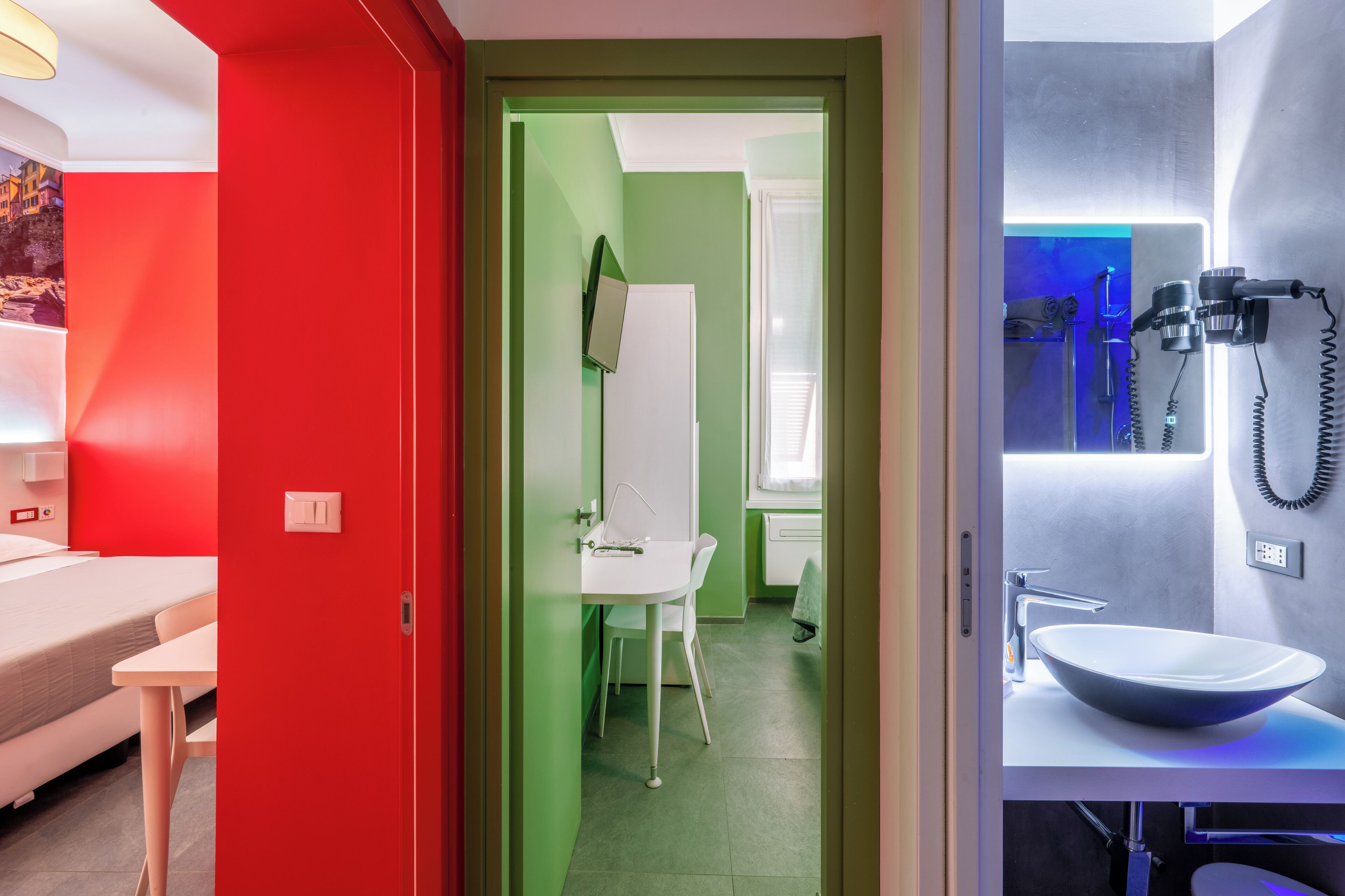 Quarto de hotel moderno com parede vermelha, cama branca, porta verde e banheiro com pia grande e iluminação azul.