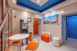 Een moderne hotel lobby met een trap, een automaat, een klok, een schilderij en een blauwe deur.