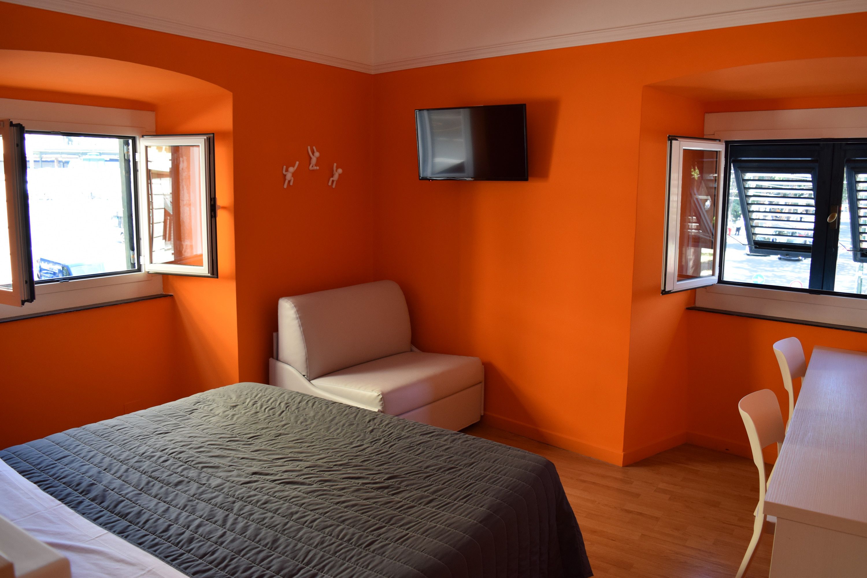 A bedroom with an orange wall, a bed, a white chair, and a TV mounted on the wall.