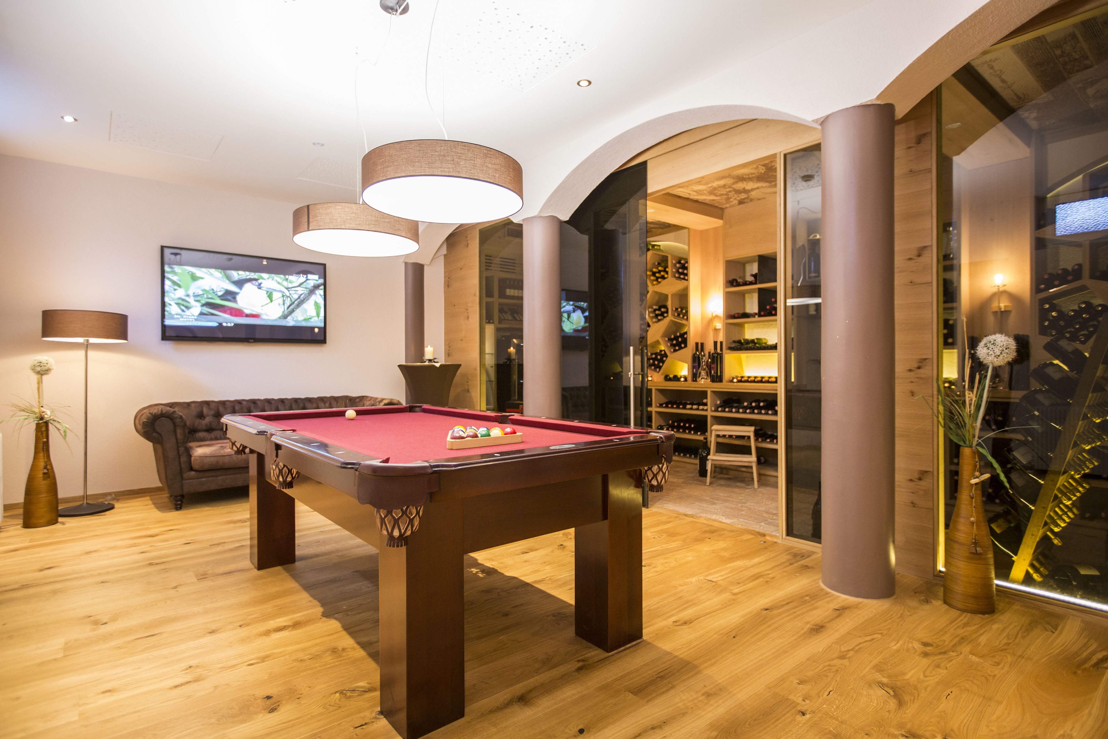 Una mesa de billar en una habitación con suelo de madera, un televisor montado en la pared y una estantería llena de libros y objetos decorativos.