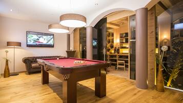 Una mesa de billar en una habitación con suelo de madera, un televisor montado en la pared y una estantería llena de libros y objetos decorativos.