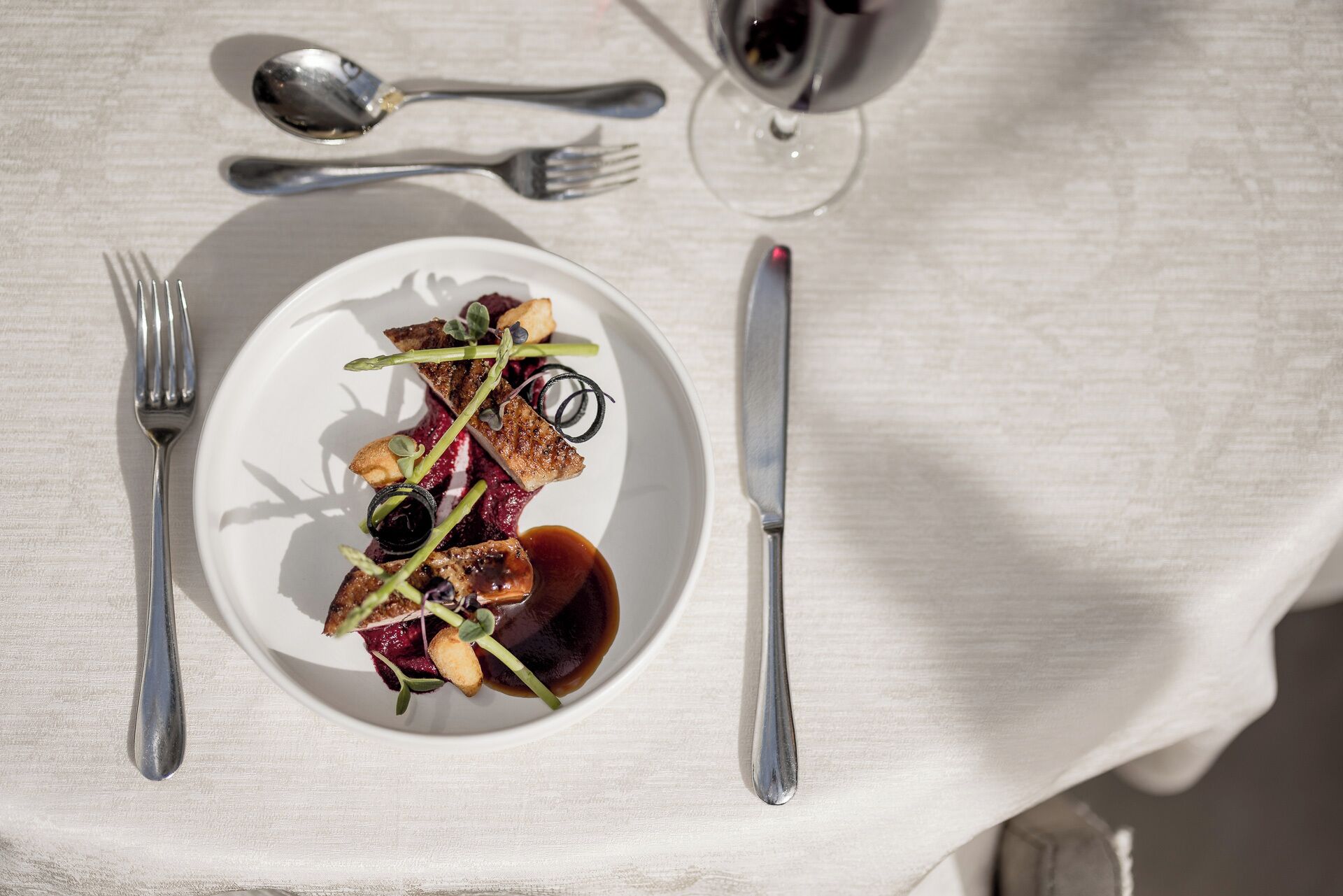 Una mesa de comedor con un plato de comida, cubiertos y una copa de vino tinto.