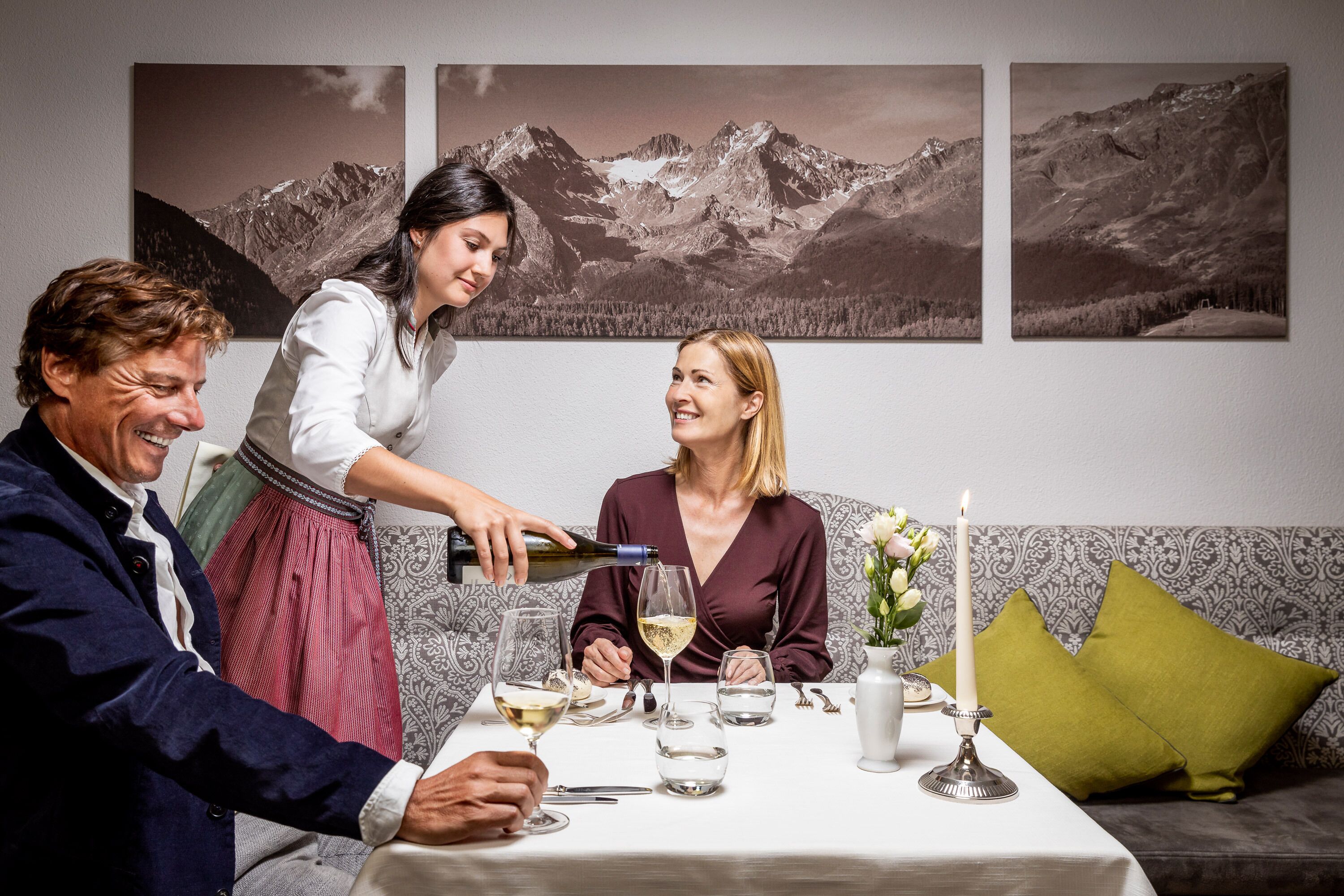 Una mesa de comedor con mantel blanco, copas de vino y una vela. Tres personas están sentadas alrededor de la mesa.