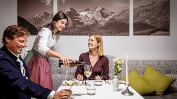 Una mesa de comedor con mantel blanco, copas de vino y una vela. Tres personas están sentadas alrededor de la mesa.