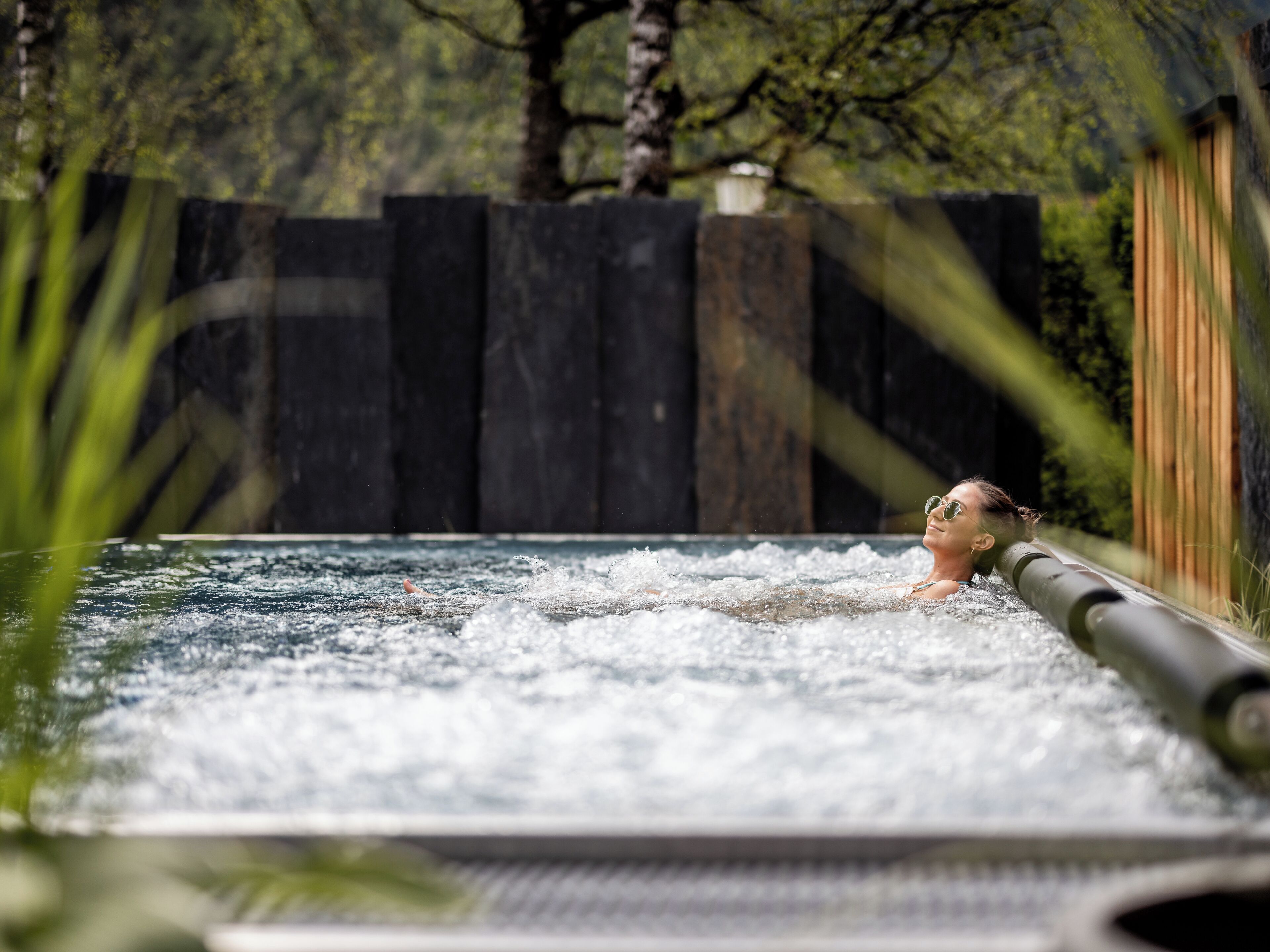 Una persona en un jacuzzi con chorros de agua, rodeada de una valla de madera y vegetación.