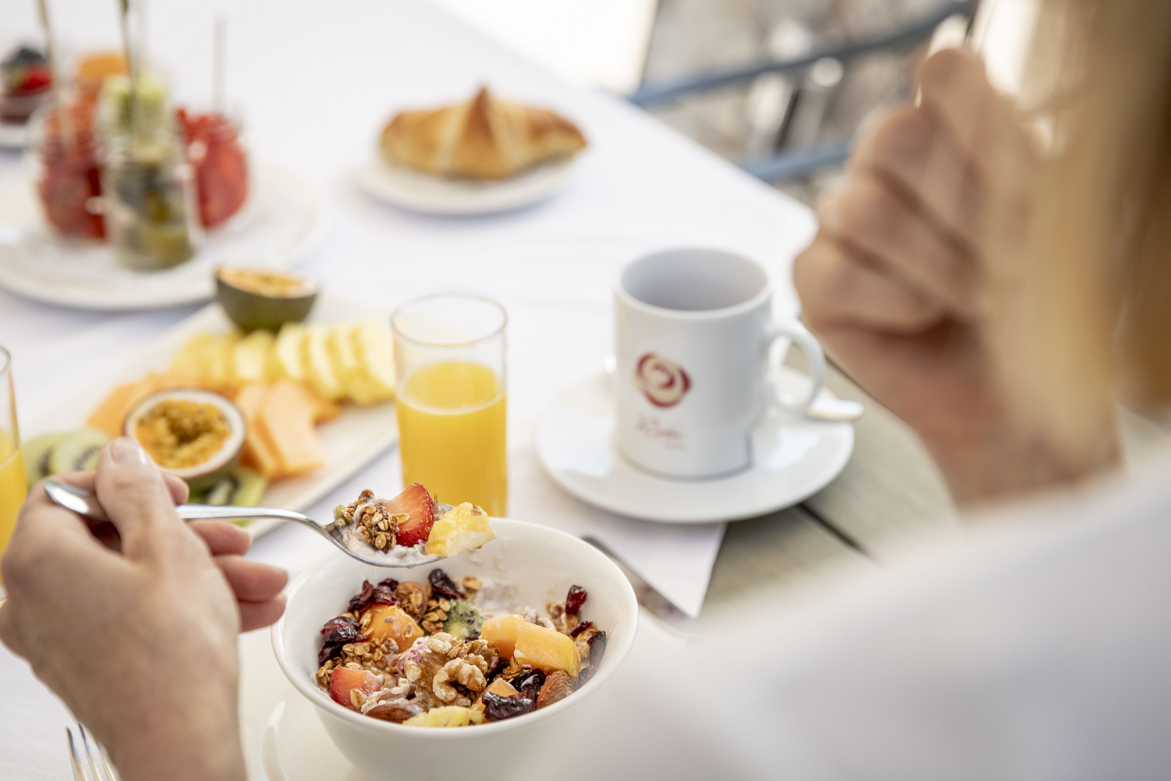 A breakfast setting with a bowl of cereal, a glass of orange juice, and a croissant on a white table.
