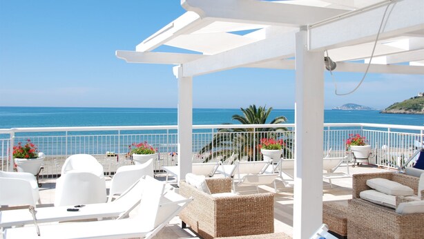 A covered outdoor seating area with white furniture and potted plants, overlooking a beach and the sea. - Hotel Villa Eleonora (Minturno)