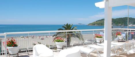 A balcony with white chairs and tables, overlooking a beach with umbrellas and people.
