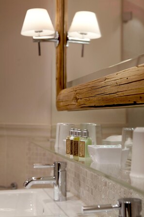 Une salle de bain avec une étagère en verre où sont rangés les articles de toilette, un miroir à cadre en bois et un lavabo blanc.