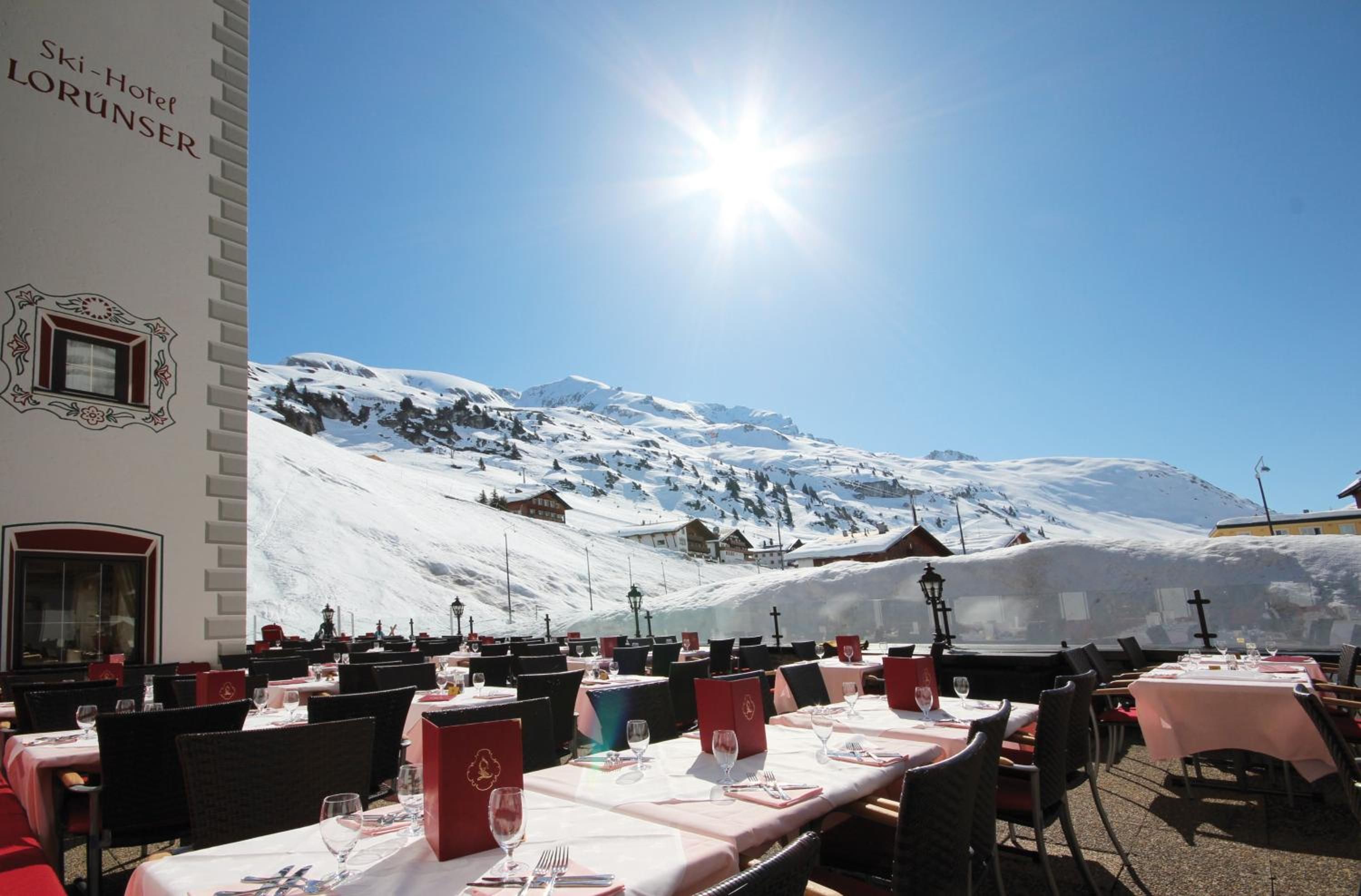 A restaurant with tables set for dining, a mountain backdrop, and a building with signage.