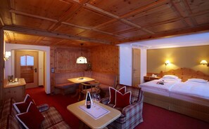 A hotel room with a wooden ceiling, a bed, a sofa, a table with a bottle and glasses, and a small dining table.