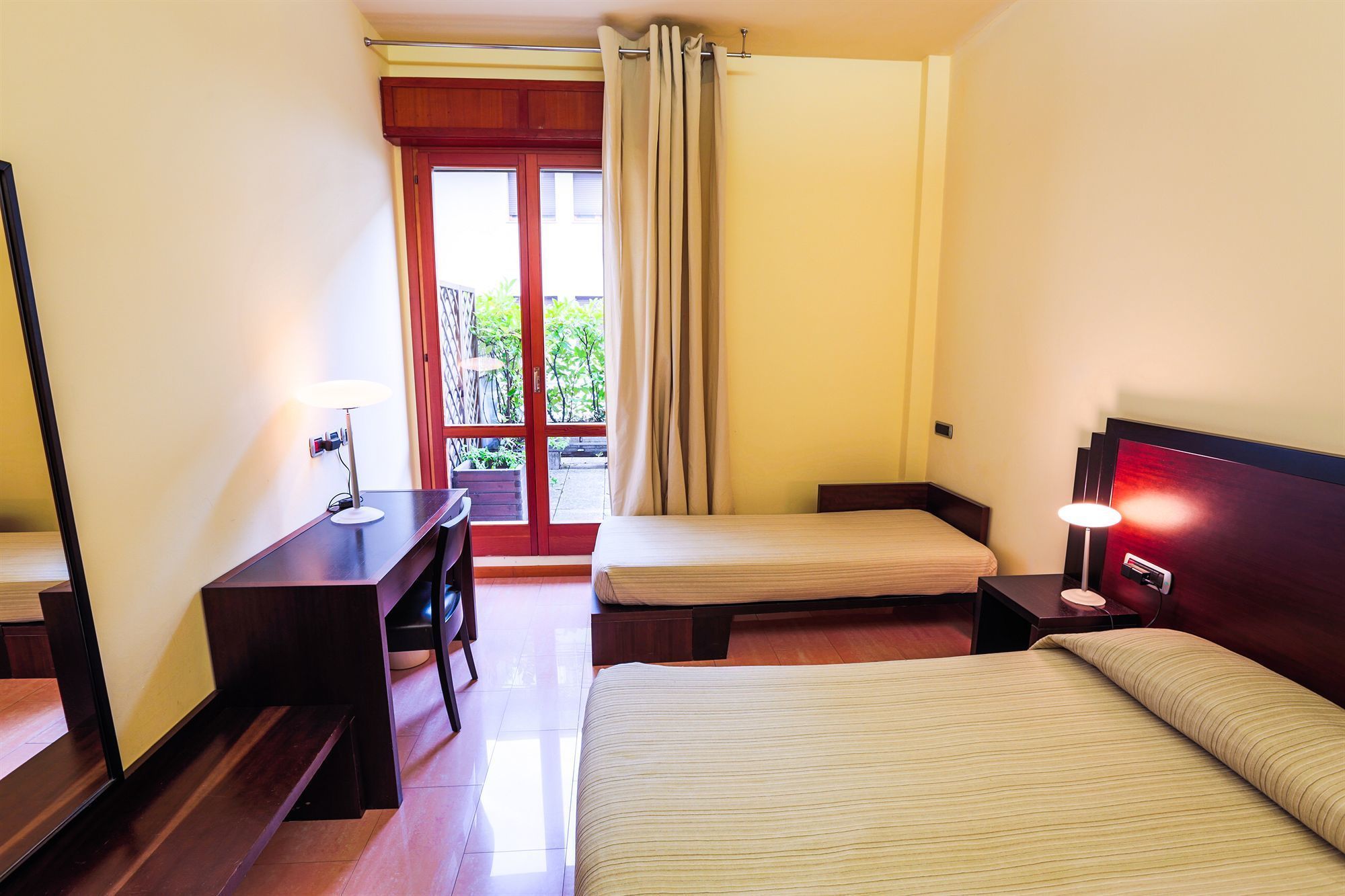 A hotel room with a bed, a desk, a chair, a nightstand, and a lamp. There is a window with curtains and a view of greenery outside.