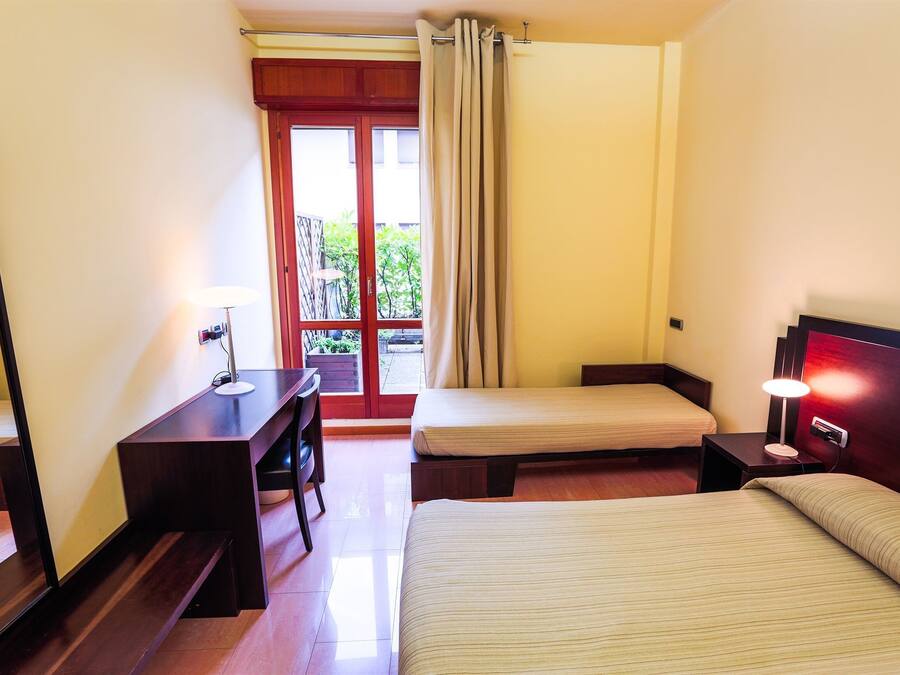 Habitación de hotel con cama, escritorio, silla, mesita de noche y lámpara. Se ve un ventanal con cortinas y vistas a la vegetación.