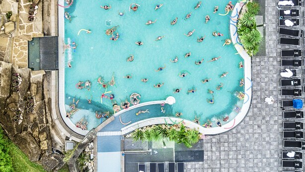 Vista aérea de una piscina con múltiples carriles, rodeada de sillas de descanso y un estacionamiento.