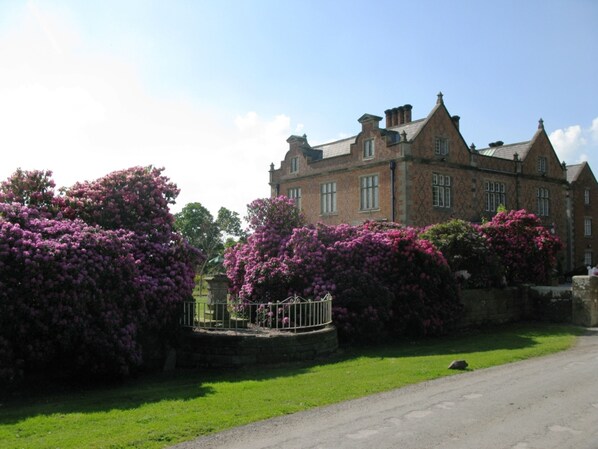 A large, historic building with a symmetrical facade, surrounded by lush greenery and flowering bushes.