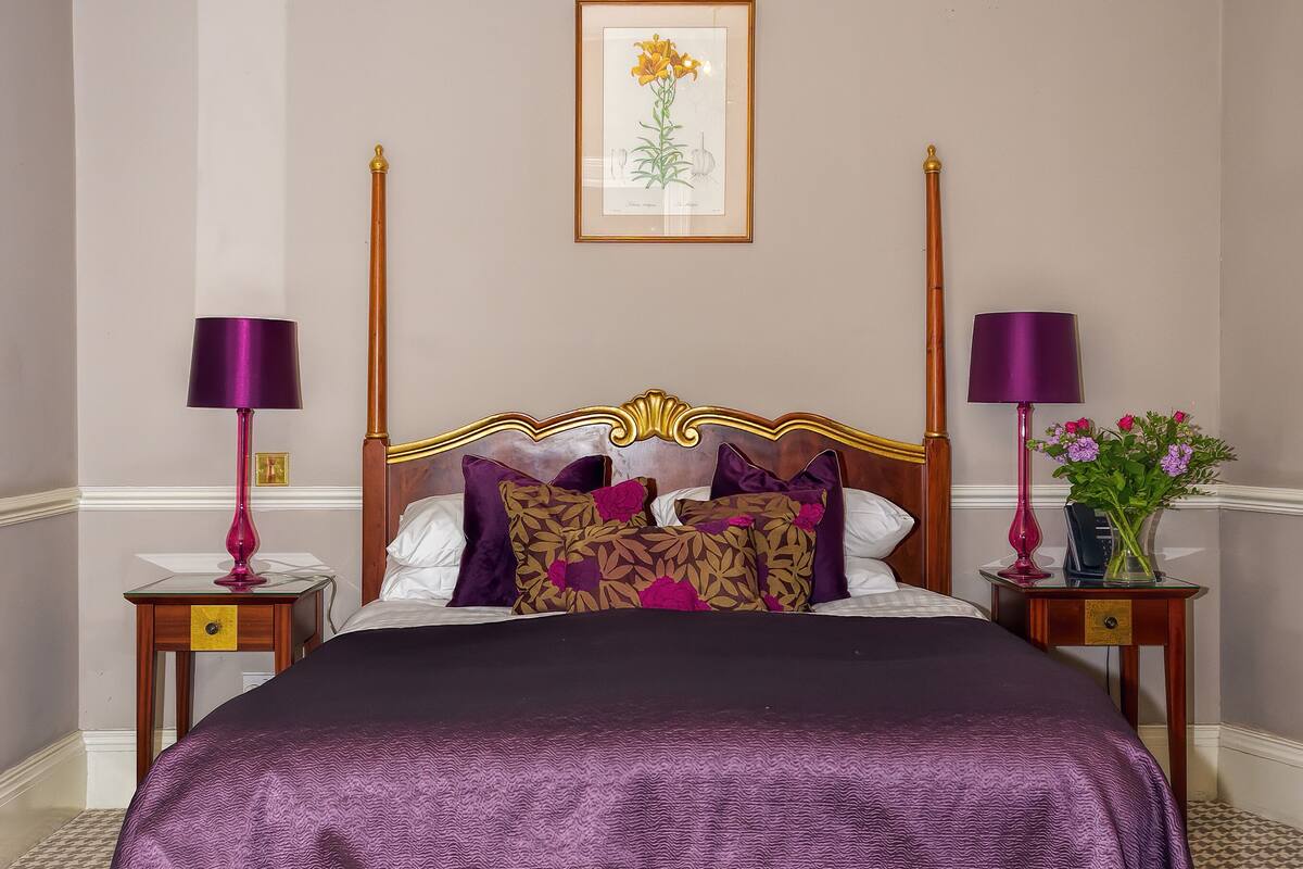 Un lit bien fait, recouvert d’une couvre-lit aux tons violets et beiges, flanqué de deux tables de chevet sur lesquelles reposent des lampes et un vase de fleurs.