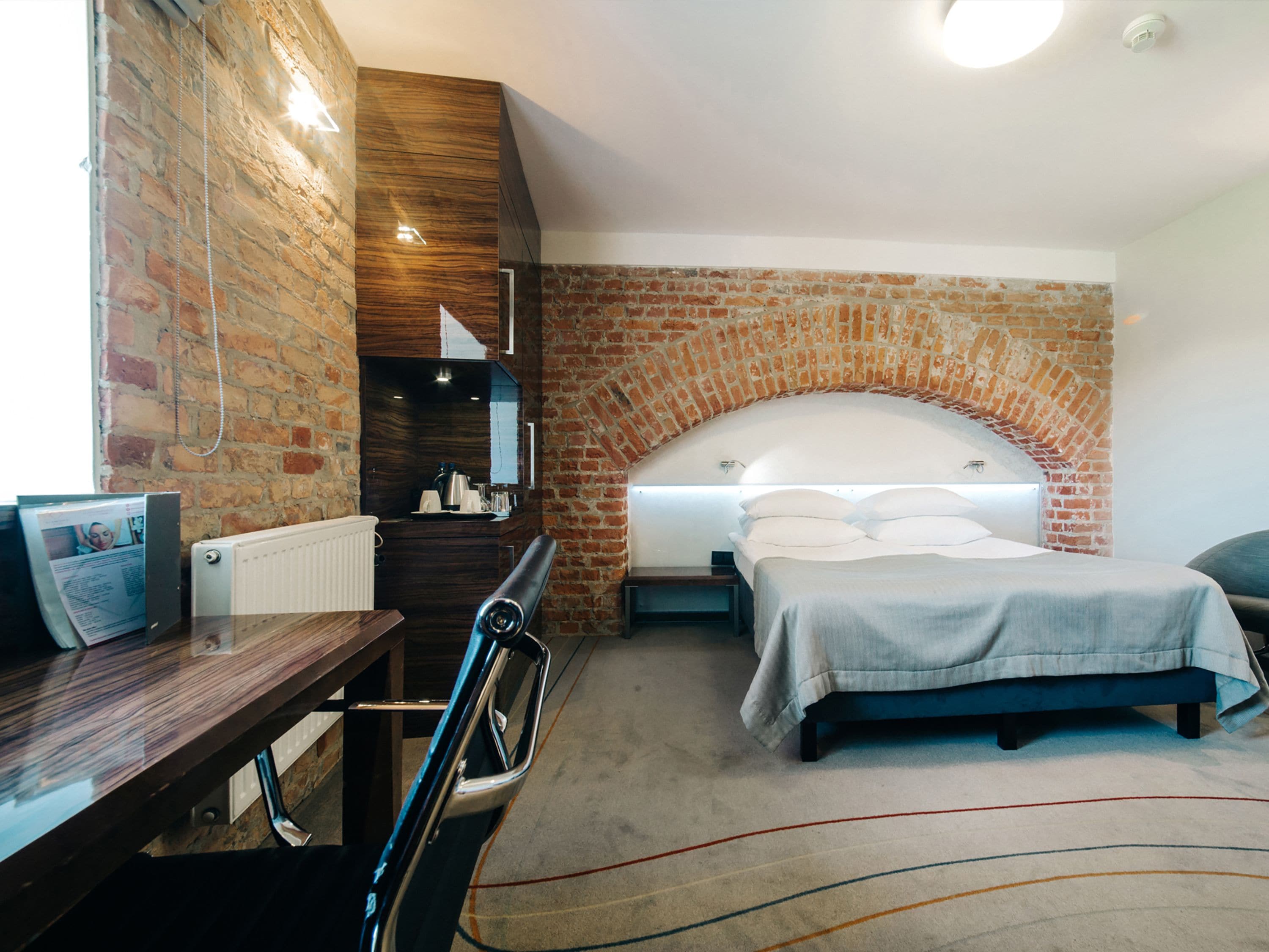 A bedroom with a brick wall, a bed with a white headboard, a desk with a chair, and a radiator.