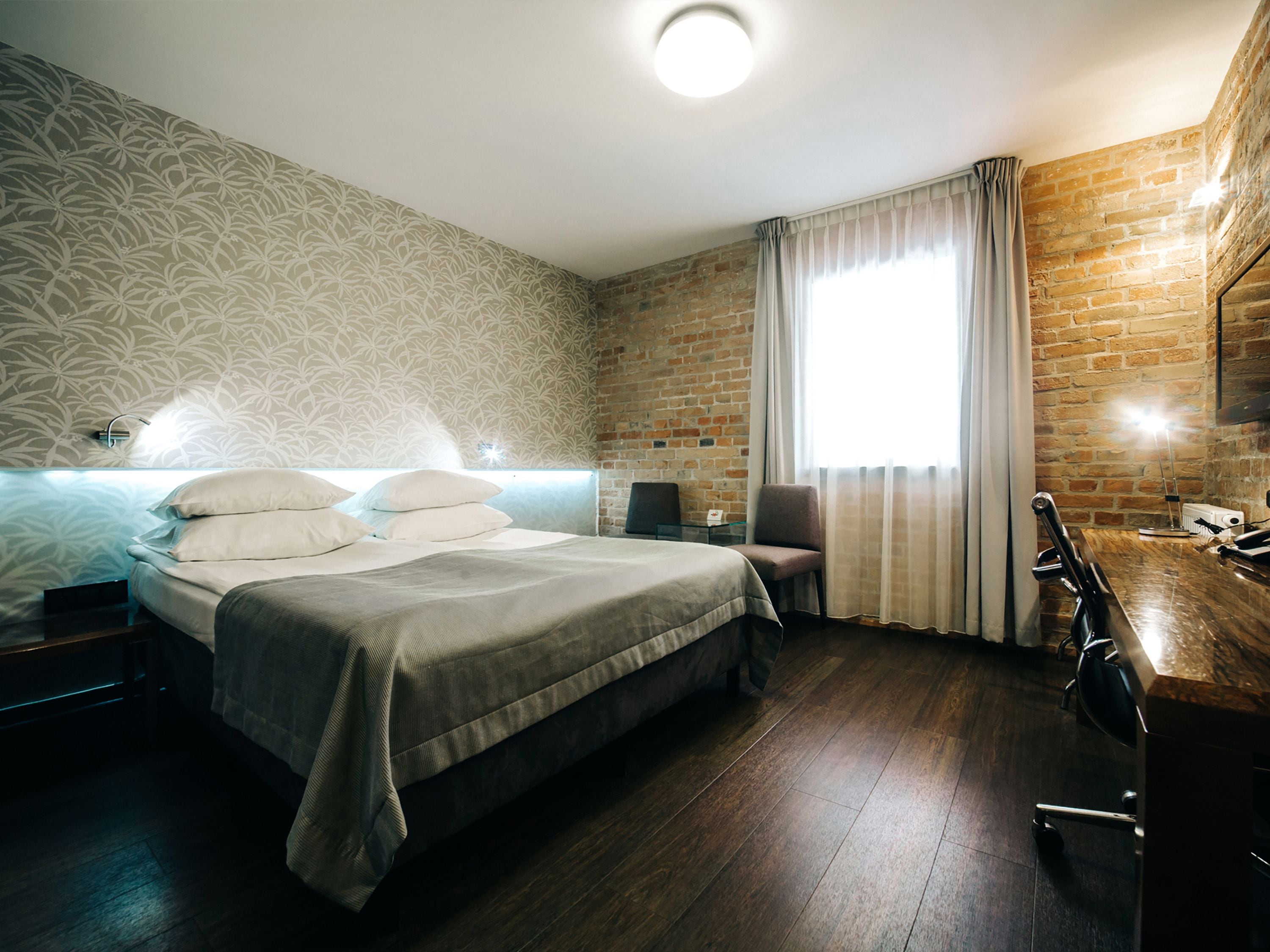 A hotel room with a large bed, a desk, and a chair. There is a brick wall and a window with curtains.