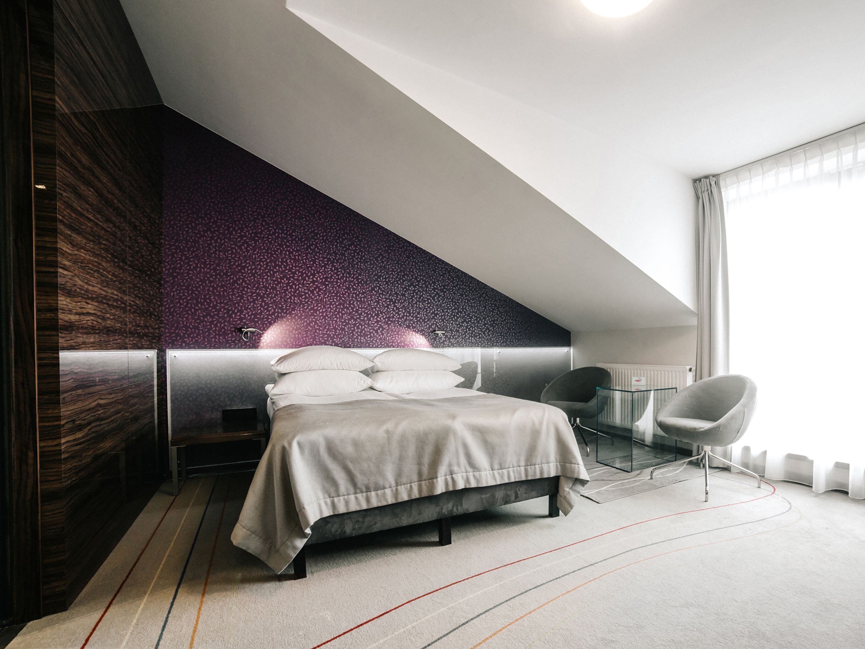 A modern bedroom with a sloped ceiling, a bed with white bedding, a glass side table, a chair, and a view of the outdoors through a window.