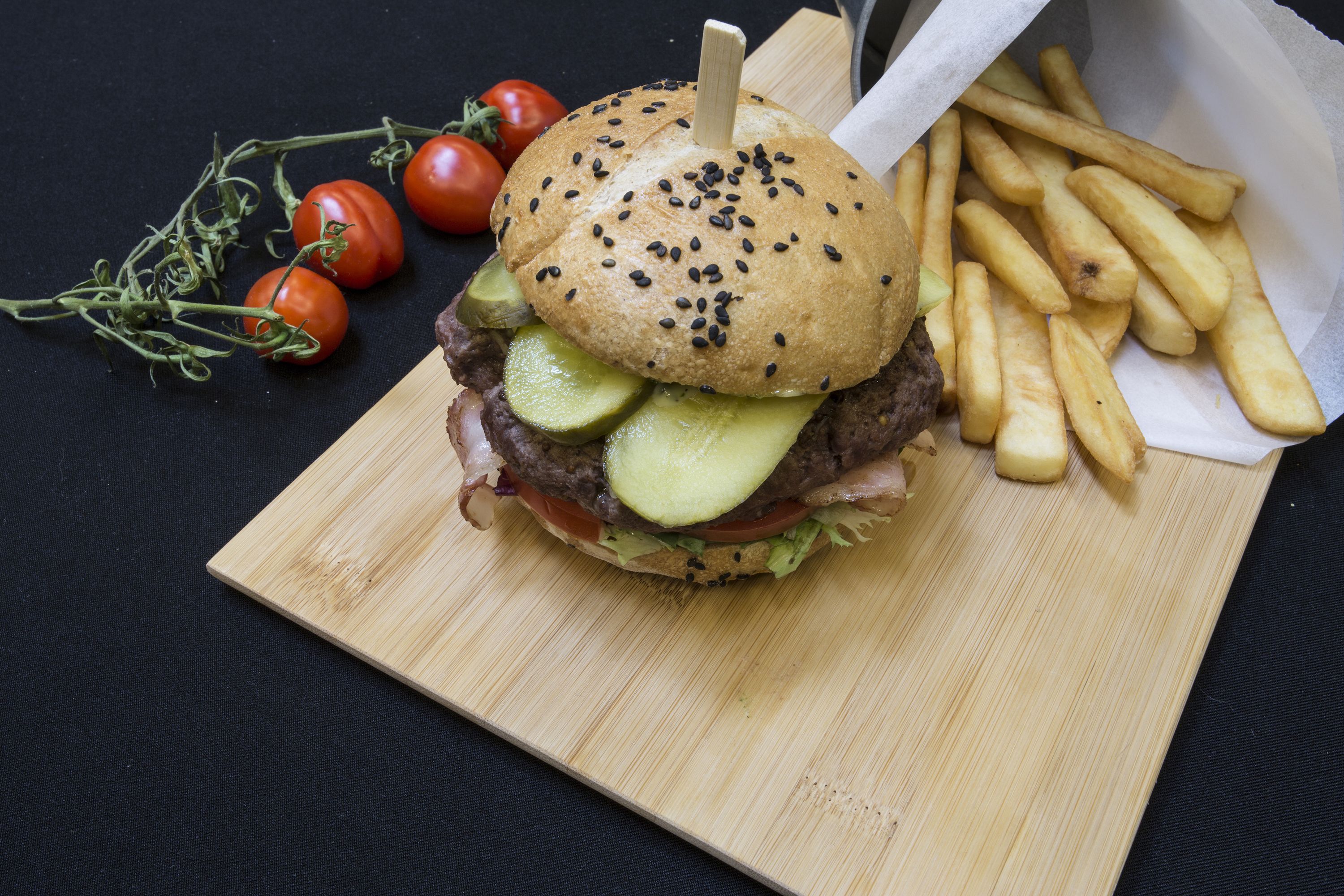Un hamburger servi dans un pain aux graines de sésame, garni de laitue, de tomates et de cornichons, accompagné de frites sur une planche en bois.