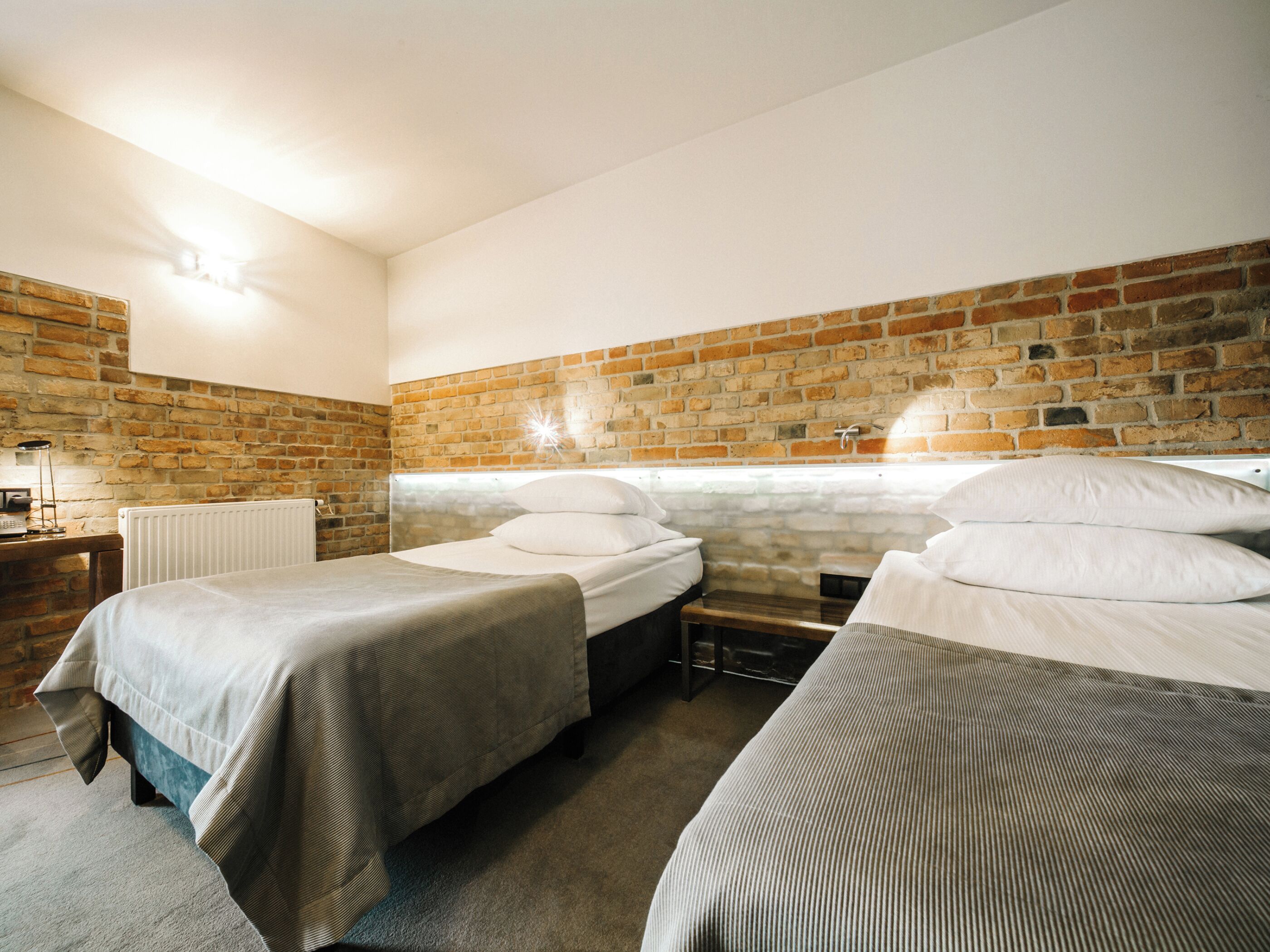 A room with two beds, a brick wall, a radiator, and a small table with a lamp.
