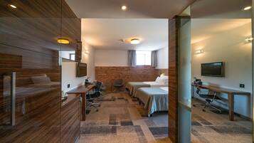 A modern hotel room with a wooden wardrobe, a desk with a chair, two beds, a TV, and a brick wall.