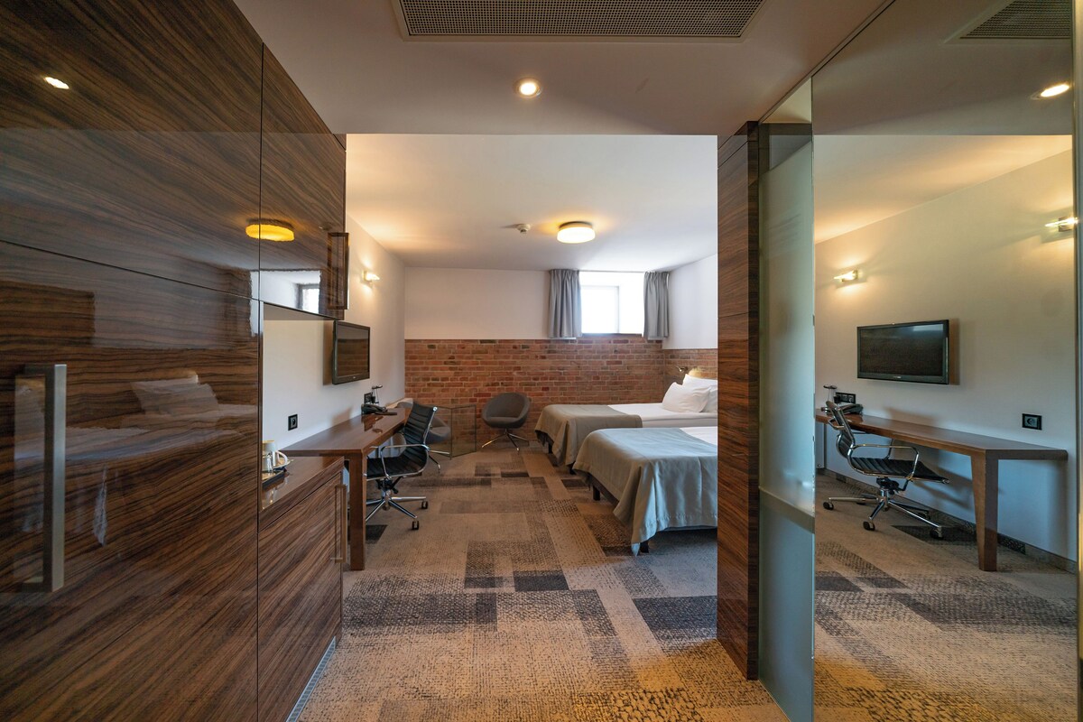 A modern hotel room with a wooden wardrobe, a desk with a chair, two beds, a TV, and a brick wall.