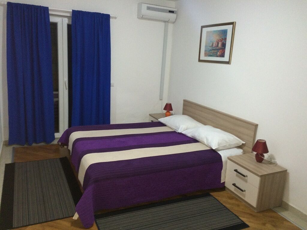 A bedroom with two beds, a wooden headboard, bedside tables with lamps, a painting on the wall, and a window with blue curtains.
