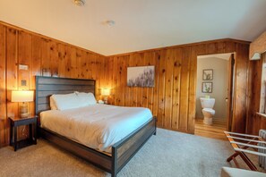 A bedroom with wooden paneling, a bed, bedside lamps, a framed picture, and a view into the bathroom.