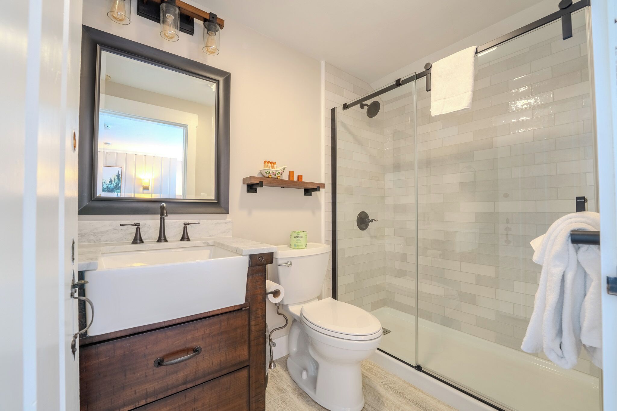 Un baño moderno con ducha envuelta en vidrio, inodoro blanco, tocador con espejo y repisas con artículos de aseo.