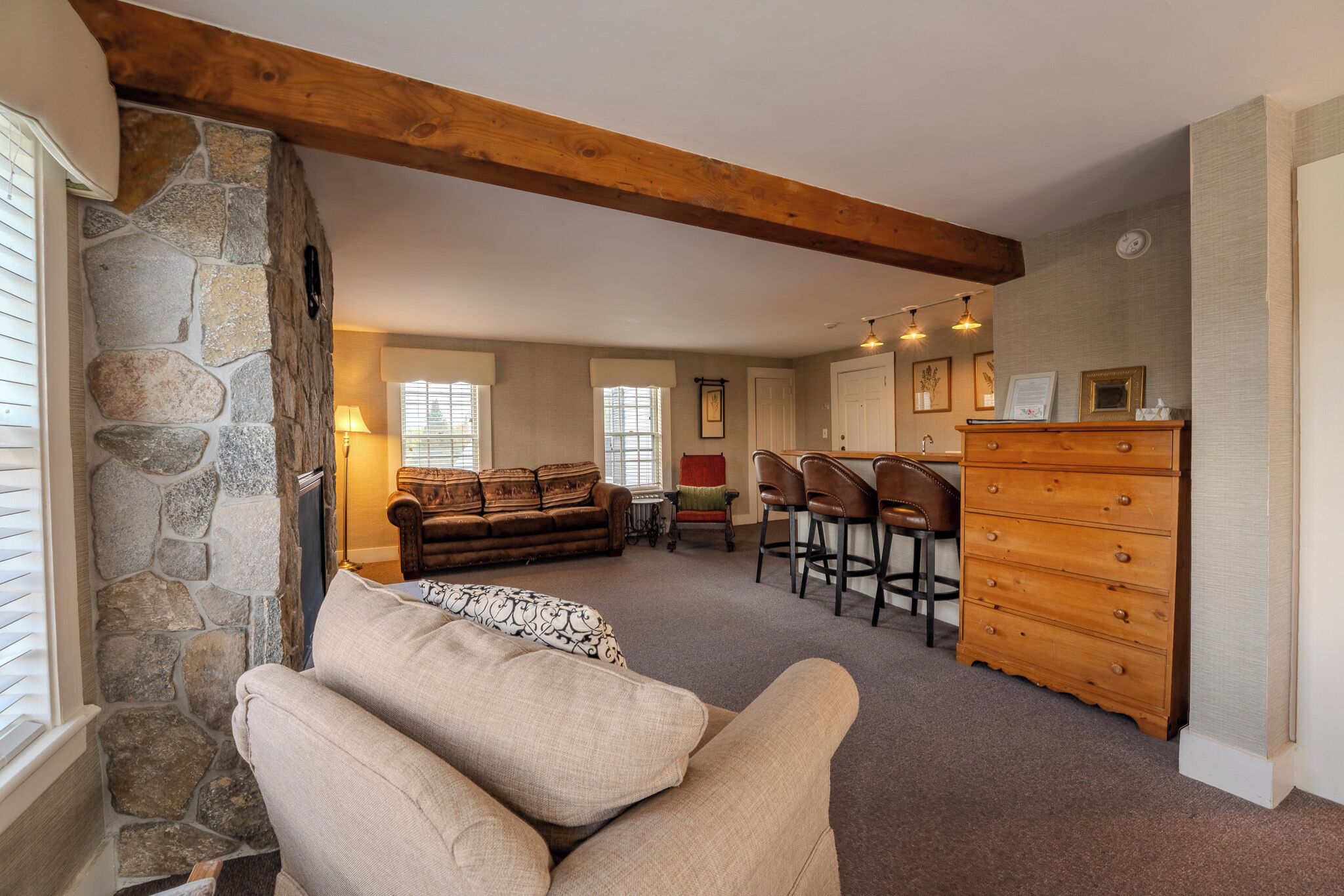 A cozy living room with a stone wall, wooden beams, a sofa, a coffee table, and a bar area with stools and a wooden dresser.
