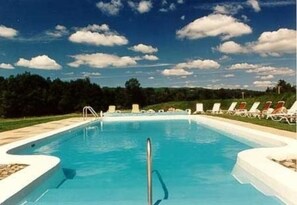 A rectangular swimming pool with a clear blue water surface, surrounded by a white pool deck. There are lounge chairs aligned along the edge of the pool.