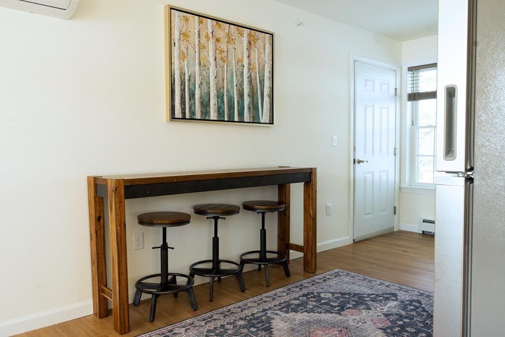 A modern kitchen with a wooden island and bar stools, a large painting above the island, and a patterned rug on the floor.